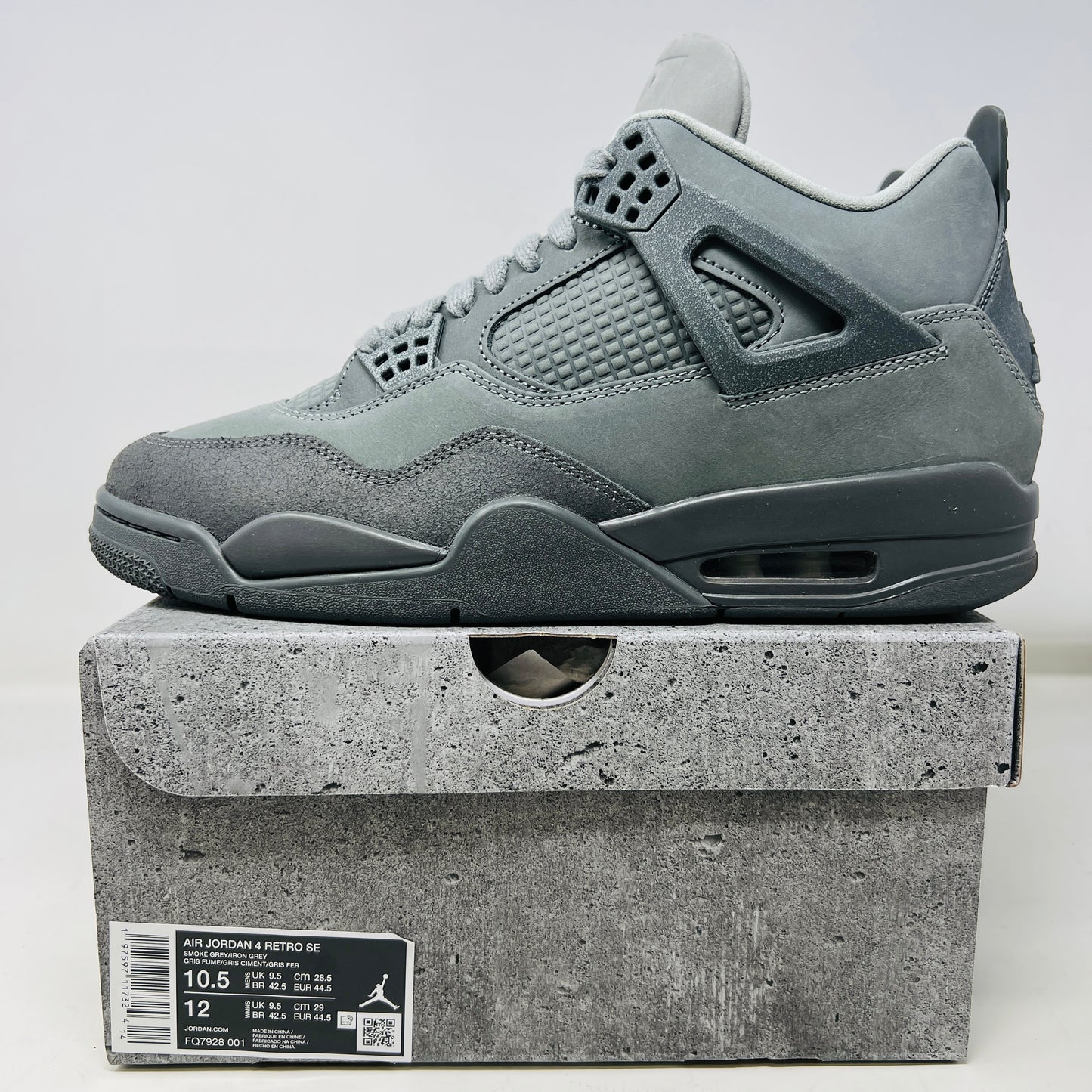 The Jordan 4 Retro SE Paris Olympics Wet Cement sneaker is displayed on its matching gray textured box, showcasing both in pristine condition. It features clean uppers with smooth and mesh textures, a visible air unit, and a size label with US, UK, and EU measurements.