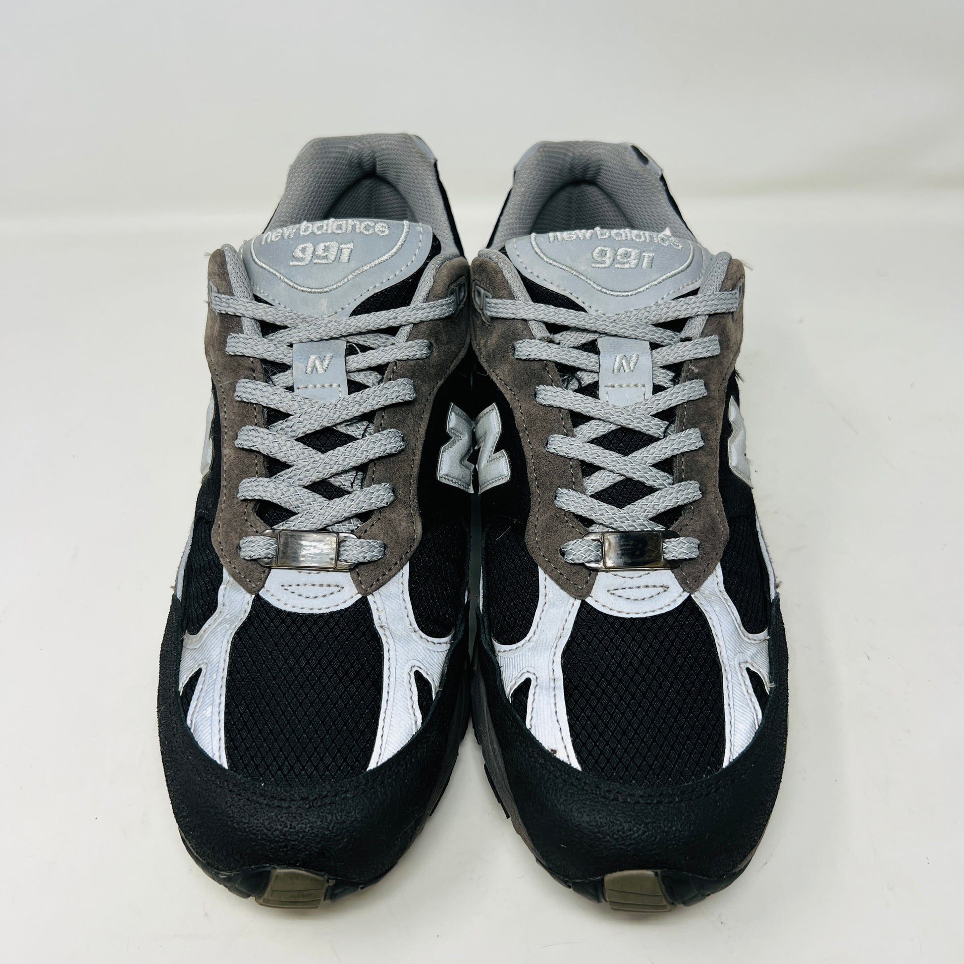 A New Balance 991 Slam Jam sneaker, in black and gray, sits on its box with tags attached. Red and black extra laces are neatly displayed in front, ready to refresh its look.