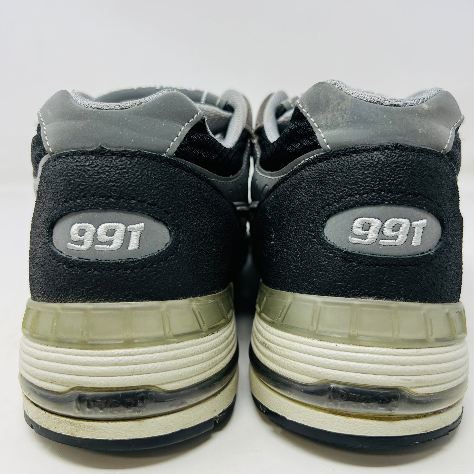 A New Balance 991 Slam Jam sneaker, in black and gray, sits on its box with tags attached. Red and black extra laces are neatly displayed in front, ready to refresh its look.