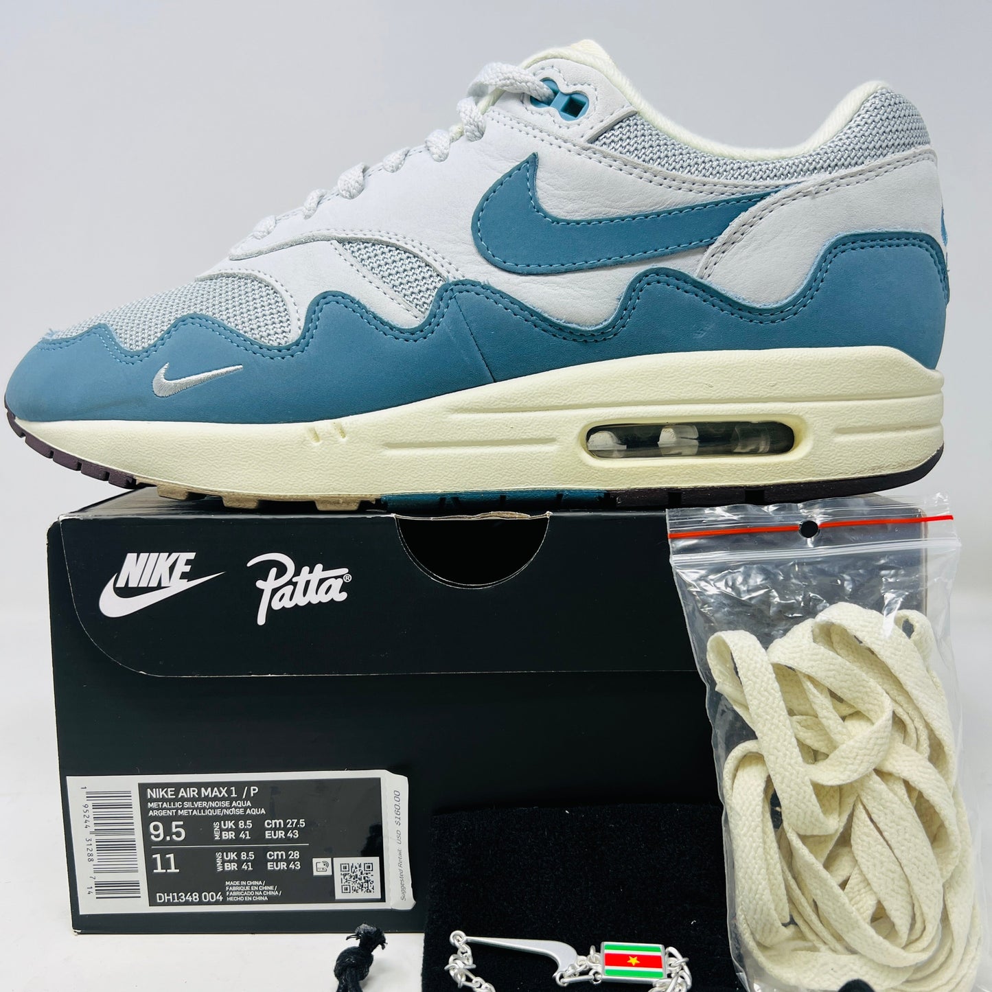The Nike Air Max 1 Patta Waves Noise Aqua features blue and white uppers and is presented on a black box. It includes a size label (US 9.5/11), extra laces in a plastic bag, and a flag-design bracelet.