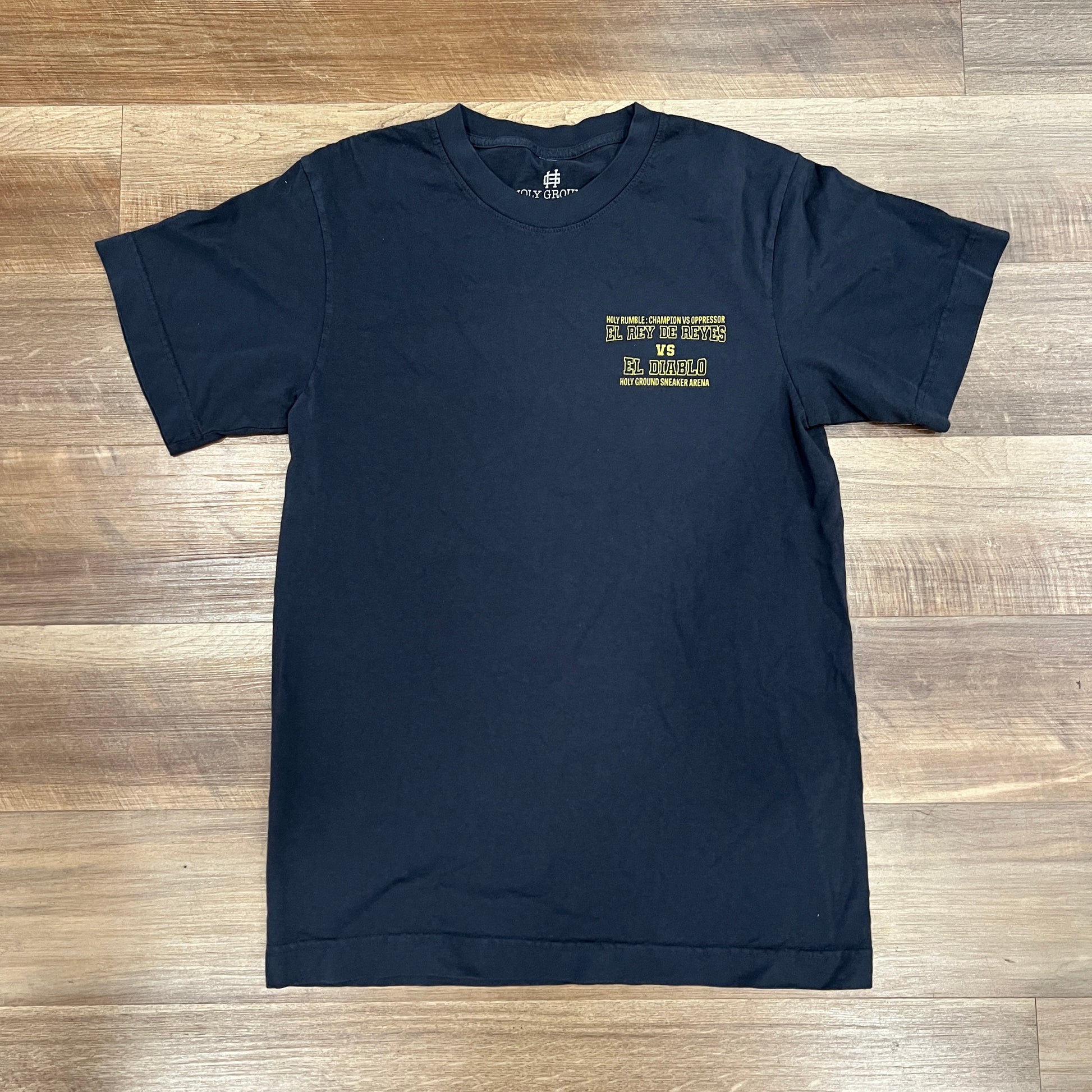 The Holy Rumble Tee by Holy Ground is a brand new black T-shirt, available in sizes S-XXL, featuring a yellow design with text and graphics on the left chest. Laid flat on a wooden floor.