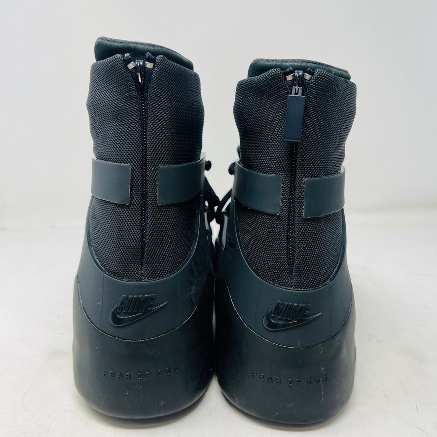The Nike Air Fear of God 1 Triple Black is a high-top, dark, minimalist sneaker with angular overlays and a textured sole. It features a high collar, matte finish, dark laces, and has a small logo near the midsole. The background is light gray with general wear signs.