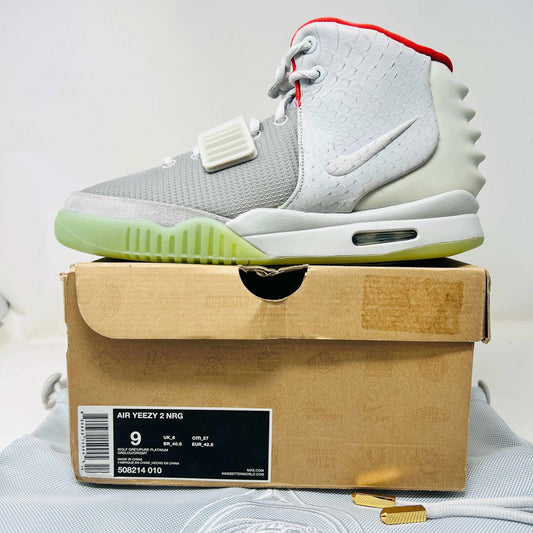 A pair of new Nike Air Yeezy 2 Pure Platinum high-tops rests on a cardboard box. The sneakers are mostly gray with a red lining, midfoot strap, and green-tinted soles. The label reads Air Yeezy 2 NRG, size 9, showcasing authentic detailing by Nike.
