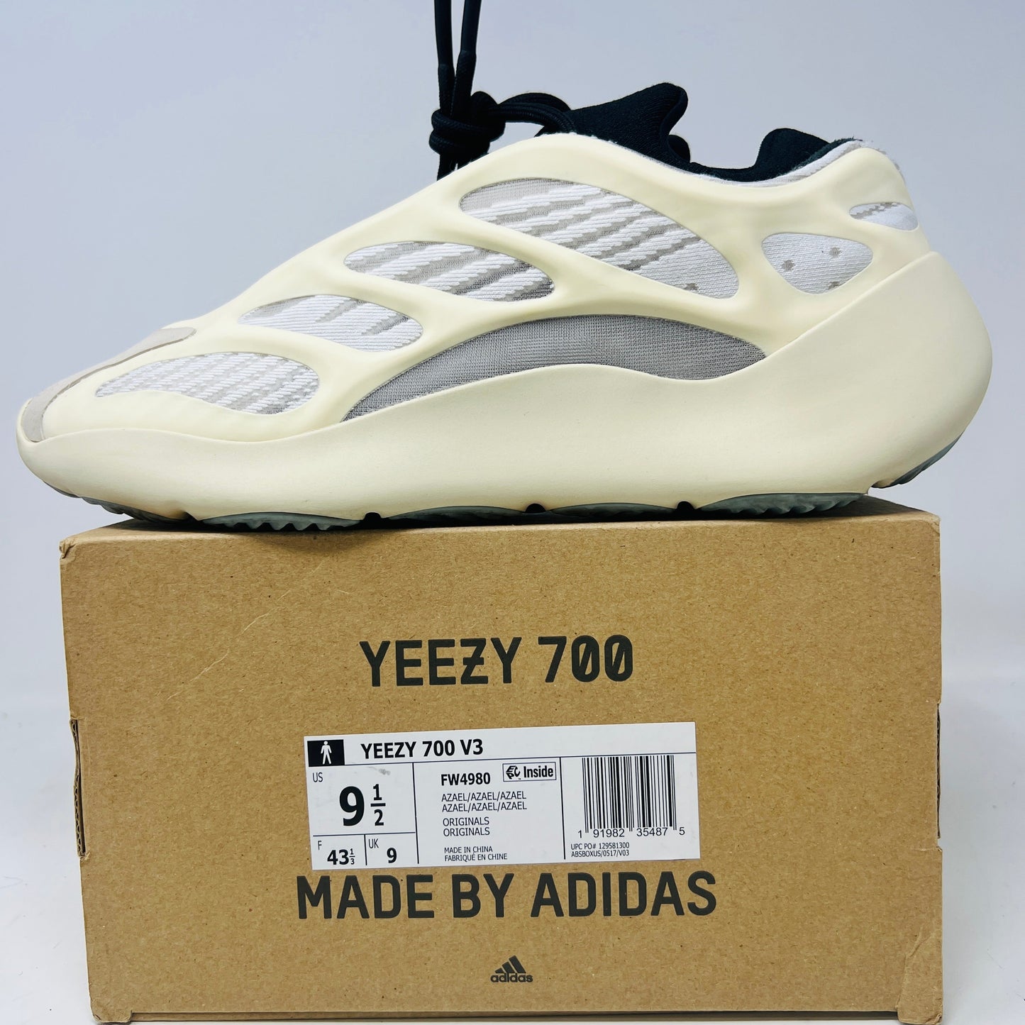 An adidas Yeezy 700 V3 Azael sneaker in a light color is displayed on a cardboard box with Adidas branding. The shoe features clean uppers, a distinctive overlay structure, and mesh inserts. Light wear is visible on minor drag areas, while the box shows size and model number details.