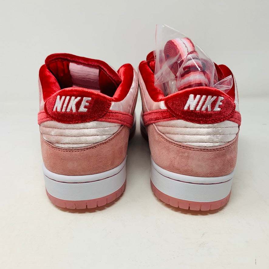 The Nike SB Dunk Low StrangeLove Skateboards, with vibrant pink and red, features a bold red swoosh, matching laces, and cartoon heart graphics on the heel. Displayed on a teal box with extra laces included, their design stands out against a plain white backdrop.