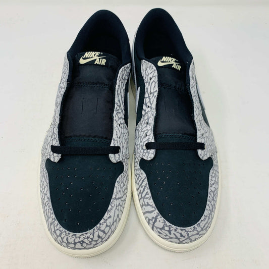 The Jordan 1 Retro Low OG Black Cement sneaker, with black and gray elephant print and a white swoosh, is showcased on a matching shoe box. A small brown box with extra laces is beside it, all set against a plain background.