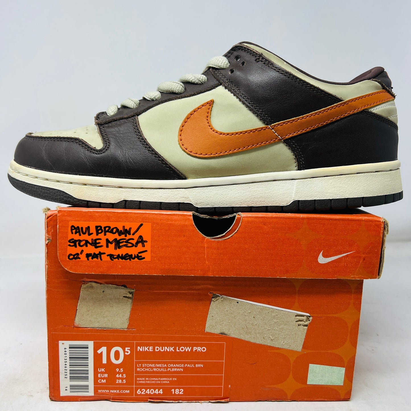 A 2002 Nike Dunk Low Light Stone Mesa Orange shoe with brown, cream, and orange hues rests on an orange box. It features an orange swoosh with a Paul Brown/Stone Mesa handwritten label. Ideal for casual wear, despite the worn box condition.