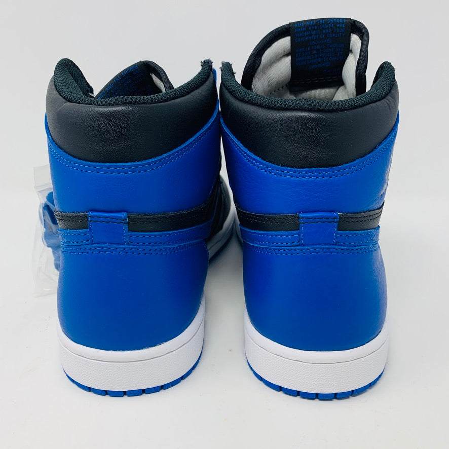 A pair of brand new black and blue Jordan 1 Royal 2017 high-tops are showcased on a black and red Nike box, with a small plastic bag holding authentic blue laces attached to one shoe.