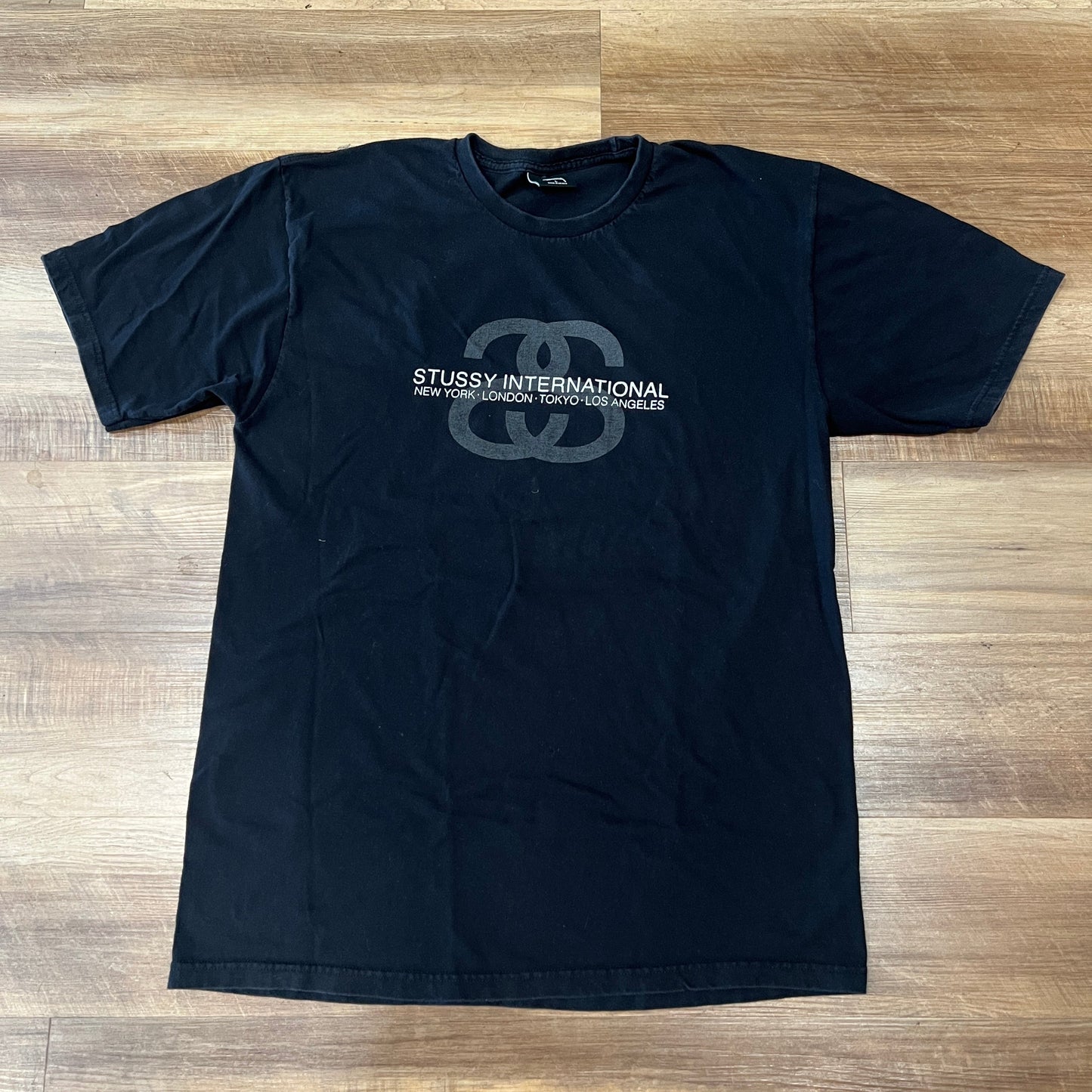 Black Stussy International Designs S Logo T-shirt laid flat on wooden floor preowned condition
