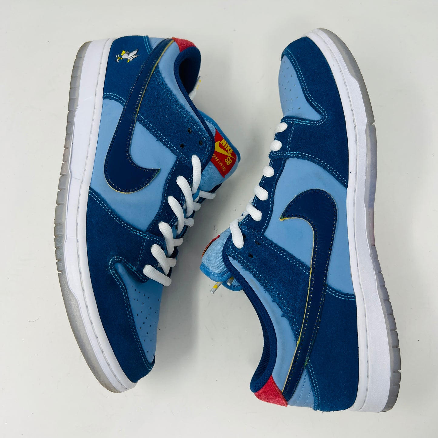 A Nike SB Dunk Low Pro Why So Sad? shoe, with blue uppers and white sole, rests on a purple box. It features an embroidered graphic on the heel, a dark blue swoosh, and extra laces in front. Price tags from 2022 are visible beside it.