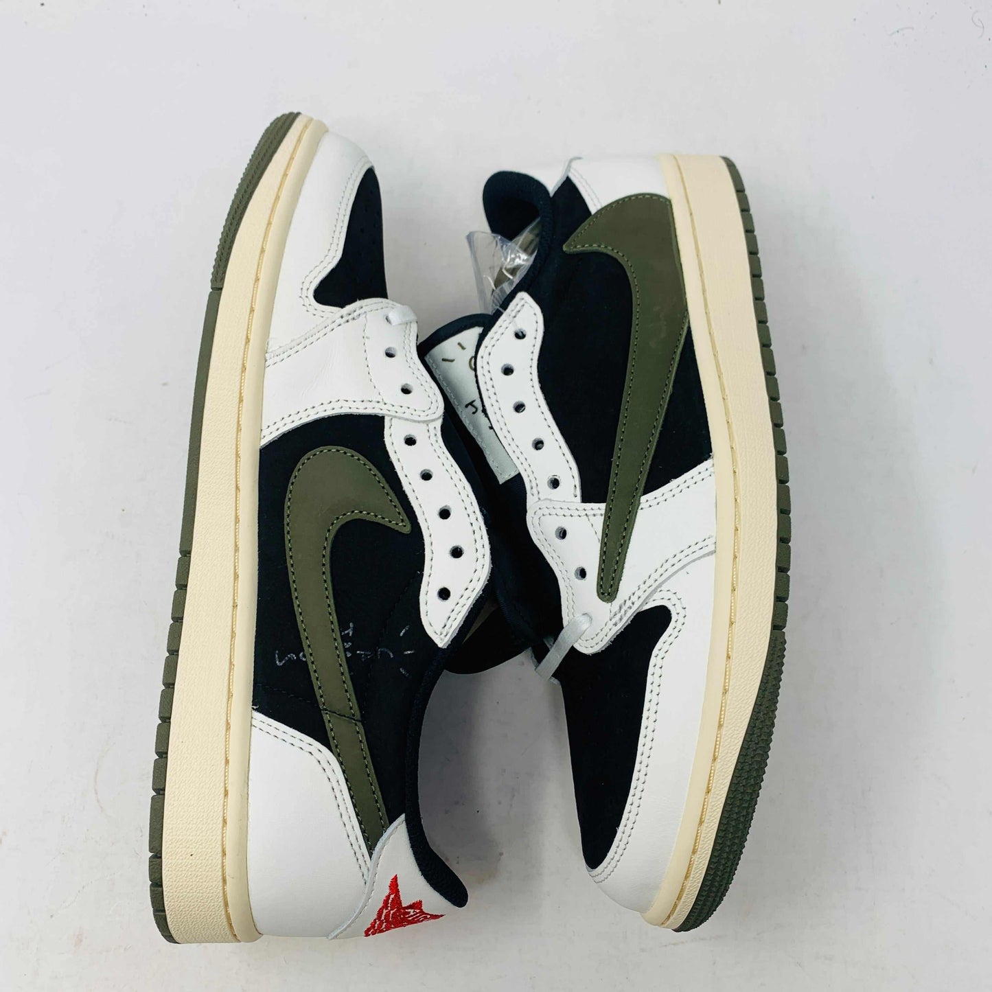 A black, white, and olive green Jordan 1 Retro Low OG SP Travis Scott Olive (W) sneaker from Nike is displayed on a matching box. This brand new shoe sports a large reversed swoosh and cream-colored sole. The box showcases the green Nike logo with 1985 Air Jordan 1 text for these authentic 2023 shoes.