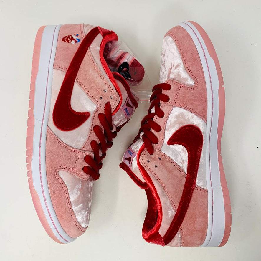 The Nike SB Dunk Low StrangeLove Skateboards, with vibrant pink and red, features a bold red swoosh, matching laces, and cartoon heart graphics on the heel. Displayed on a teal box with extra laces included, their design stands out against a plain white backdrop.