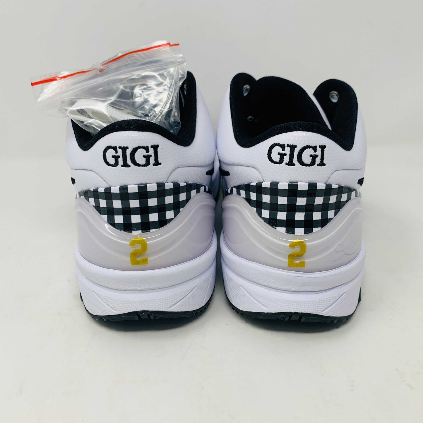 The Nike Kobe 4 Protro Mambacita Gigi shoe, featuring a black swoosh, black accents, and a checkered heel pattern, is displayed on a white box with a gold Nike logo. The box also includes extra laces.
