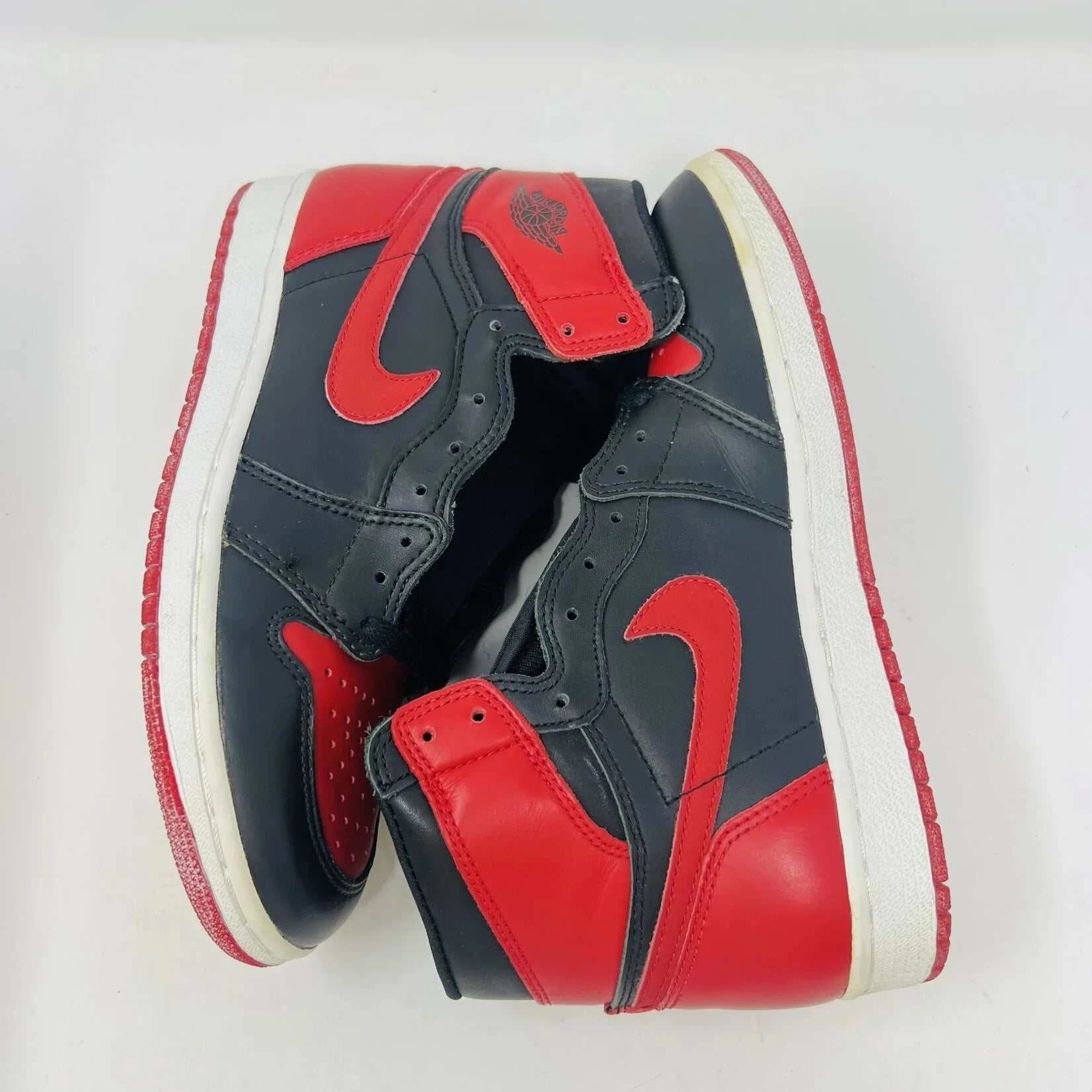A pair of brand new Jordan 1 Retro Bred (1994) sneakers is displayed on a box, featuring the iconic Nike swoosh and wings logo. A black-and-white photo of the shoes is behind them. The box has various sneaker-related graphics, emphasizing their authenticity.