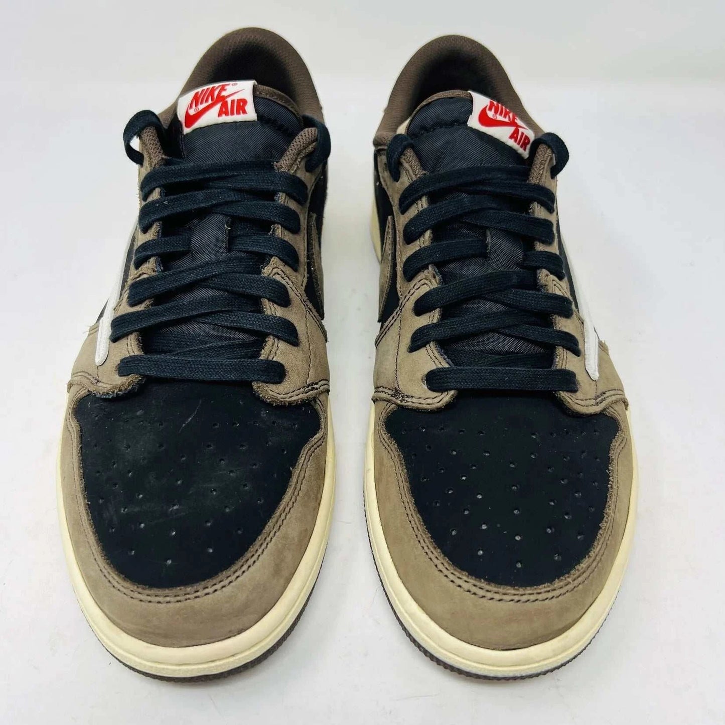 The Jordan 1 Retro Low OG SP Travis Scott Mocha sneakers, released in 2019 and featuring brown, black, and white tones, are displayed on a Nike box. Two extra lace sets, one red and one pink, are included in plastic bags beside the box.