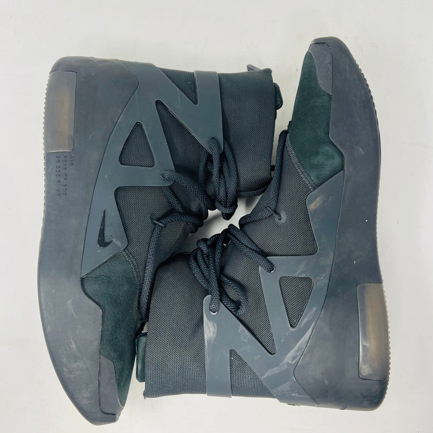 The Nike Air Fear of God 1 Triple Black is a high-top, dark, minimalist sneaker with angular overlays and a textured sole. It features a high collar, matte finish, dark laces, and has a small logo near the midsole. The background is light gray with general wear signs.
