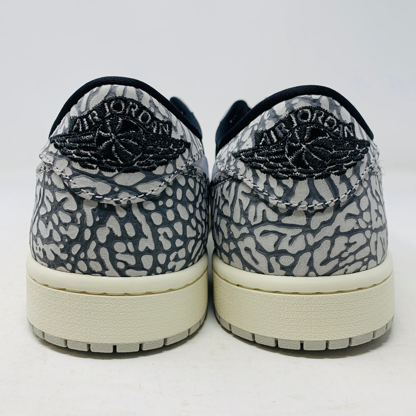 The Jordan 1 Retro Low OG Black Cement sneaker, with black and gray elephant print and a white swoosh, is showcased on a matching shoe box. A small brown box with extra laces is beside it, all set against a plain background.