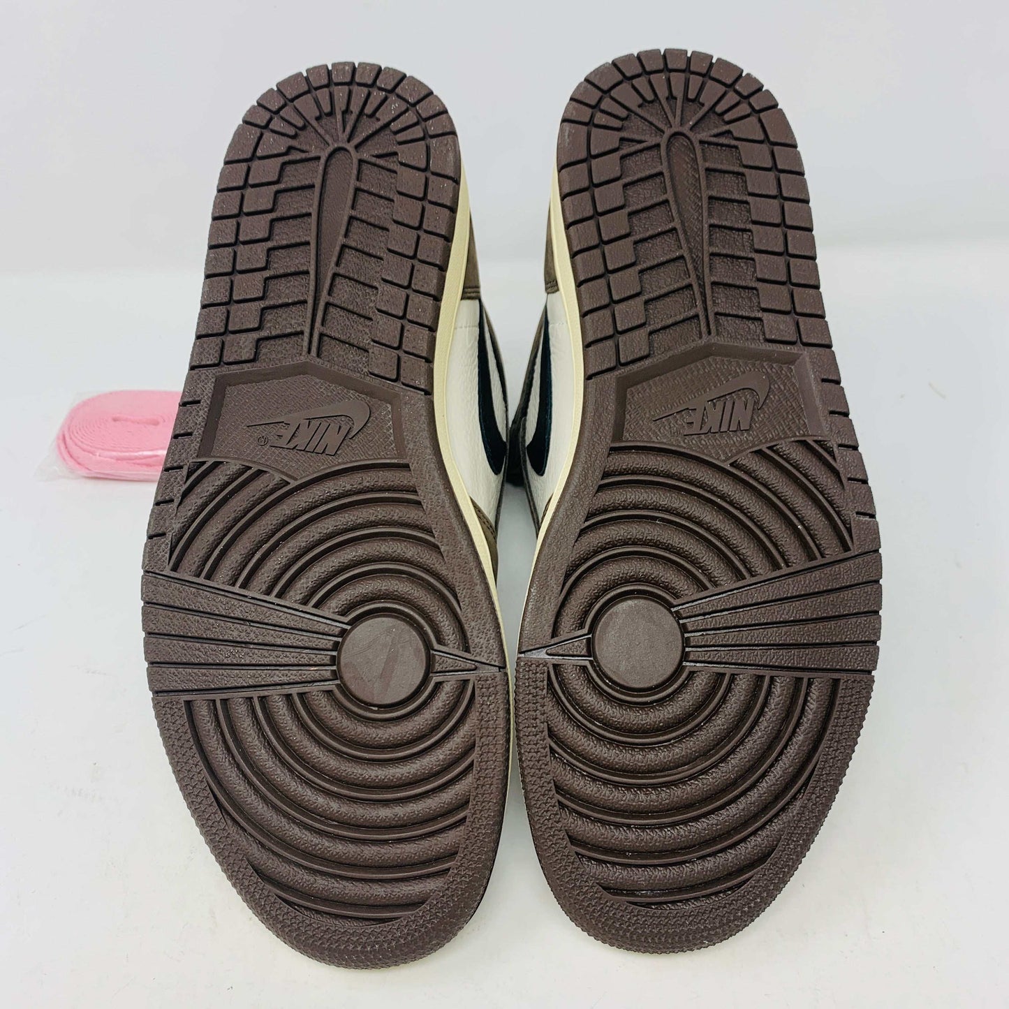 A pair of authentic Jordan 1 High Travis Scott Mocha sneakers in brown, white, and black is displayed on an orange and brown Nike shoebox. These feature a distinctive reversed black Swoosh and come with extra laces for versatile styling.