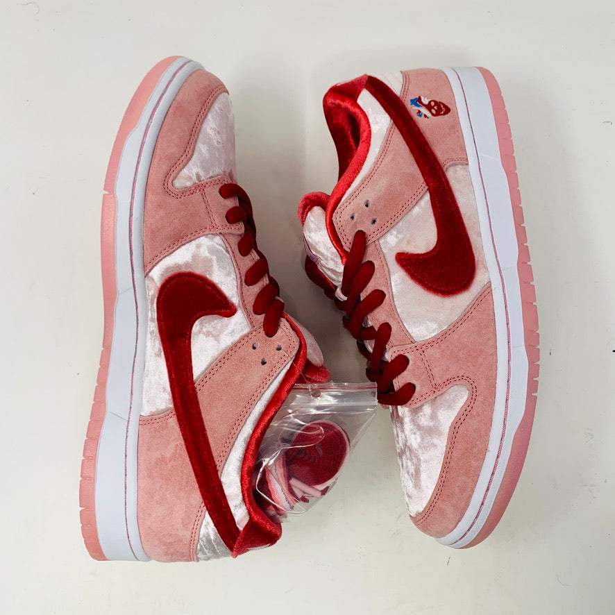 The Nike SB Dunk Low StrangeLove Skateboards, with vibrant pink and red, features a bold red swoosh, matching laces, and cartoon heart graphics on the heel. Displayed on a teal box with extra laces included, their design stands out against a plain white backdrop.