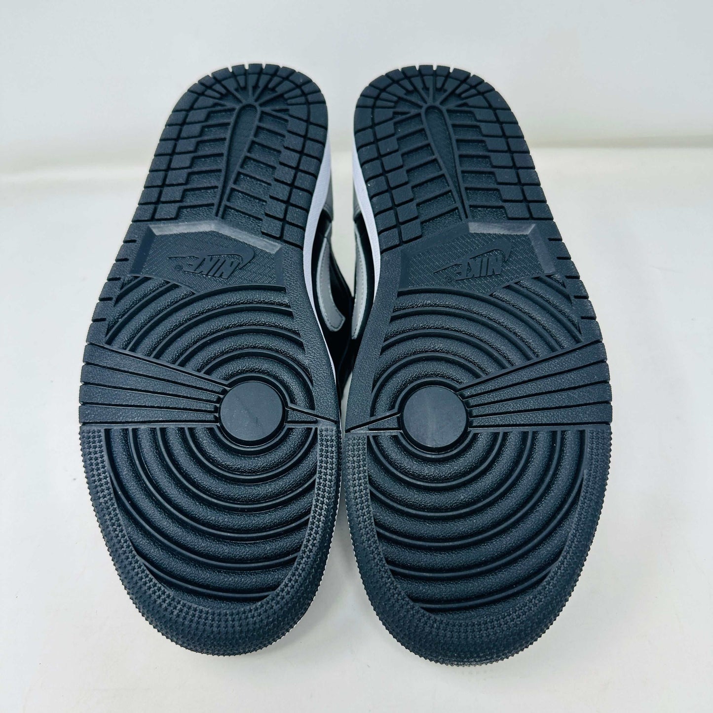 A pair of Jordan 1 Low OG Shadow (2024) sneakers, in black and gray, rests atop a gray shoe box with a large black Nike logo. Beside the box is a small brown cardboard package featuring a cutout revealing shoelaces inside.