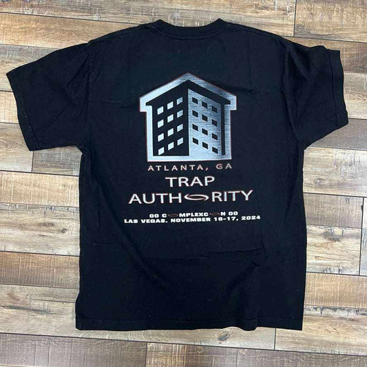 The Ken x Playboi Carti Opium Tee Black from Holy Ground Sneaker Shop lies on a wooden floor, featuring a bold building graphic and Atlanta, GA Trap Authority text. Below, it highlights ComplexCon Las Vegas, November 16-17, 2024. A must-have for any Playboi Carti fan.