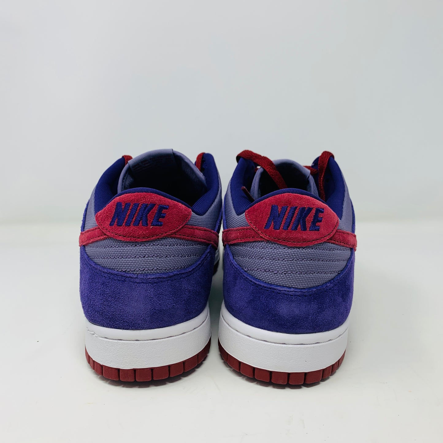 A pair of Nike Dunk Low Plum sneakers, in purple and maroon, rests on a red shoe box featuring a white Nike logo on a plain background. The shoes showcase a red swoosh and white midsoles, confirming their authenticity from Nike.