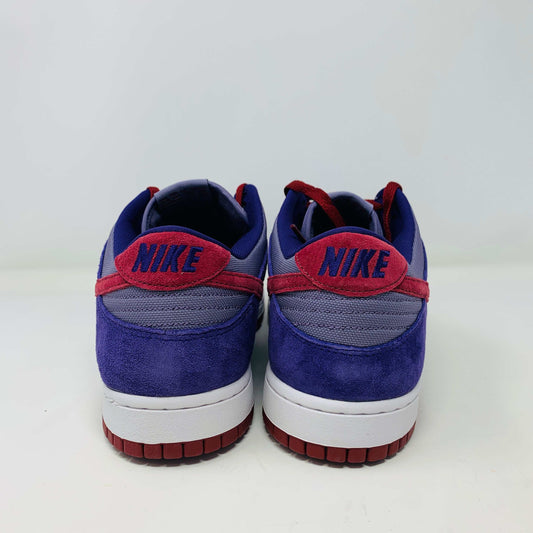A pair of Nike Dunk Low Plum sneakers, in purple and maroon, rests on a red shoe box featuring a white Nike logo on a plain background. The shoes showcase a red swoosh and white midsoles, confirming their authenticity from Nike.