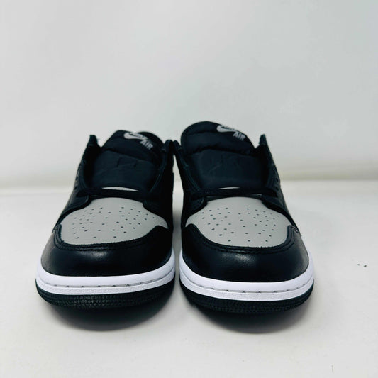 A pair of Jordan 1 Low OG Shadow (2024) sneakers, in black and gray, rests atop a gray shoe box with a large black Nike logo. Beside the box is a small brown cardboard package featuring a cutout revealing shoelaces inside.