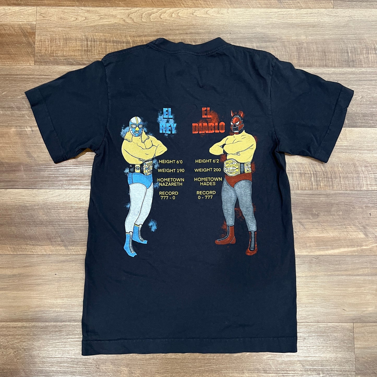 The Holy Rumble Tee by Holy Ground is a brand new black T-shirt, available in sizes S-XXL, featuring a yellow design with text and graphics on the left chest. Laid flat on a wooden floor.