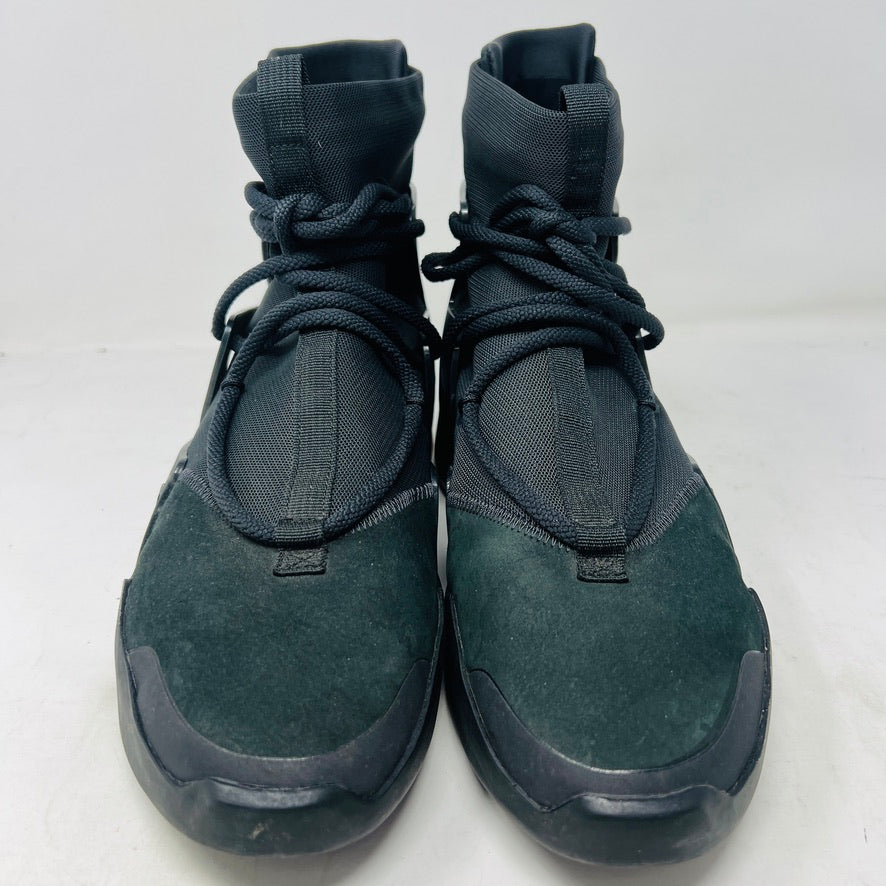 The Nike Air Fear of God 1 Triple Black is a high-top, dark, minimalist sneaker with angular overlays and a textured sole. It features a high collar, matte finish, dark laces, and has a small logo near the midsole. The background is light gray with general wear signs.