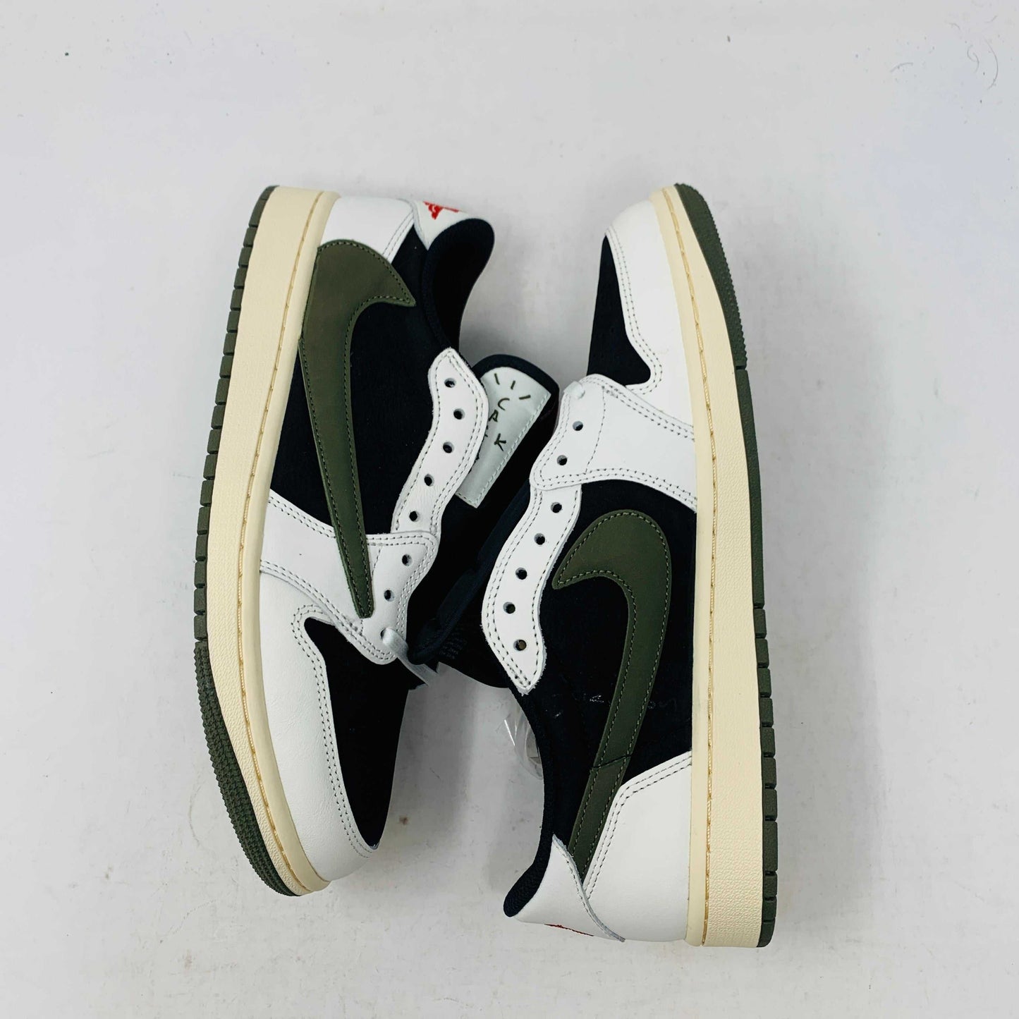 A black, white, and olive green Jordan 1 Retro Low OG SP Travis Scott Olive (W) sneaker from Nike is displayed on a matching box. This brand new shoe sports a large reversed swoosh and cream-colored sole. The box showcases the green Nike logo with 1985 Air Jordan 1 text for these authentic 2023 shoes.
