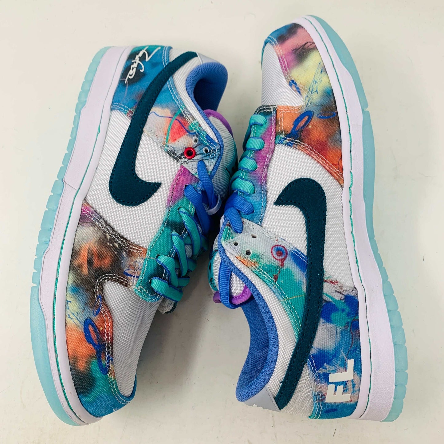 The Nike SB Dunk Low Futura Laboratories Bleached Aqua sneakers showcase a vibrant abstract design in blue, pink, orange, and turquoise. Teal swooshes and light blue soles complement the purple laces and FL branding on a pristine white surface.