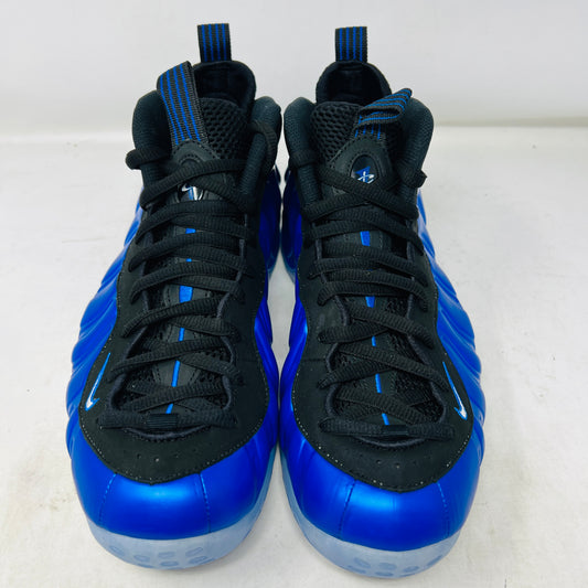 A brand-new Nike Air Foamposite One International Blue sneaker, featuring a striking blue upper with black accents, rests on a sleek black Nike shoebox. It has black mesh laces, a translucent sole with carbon fiber plate, and an iconic side swoosh.
