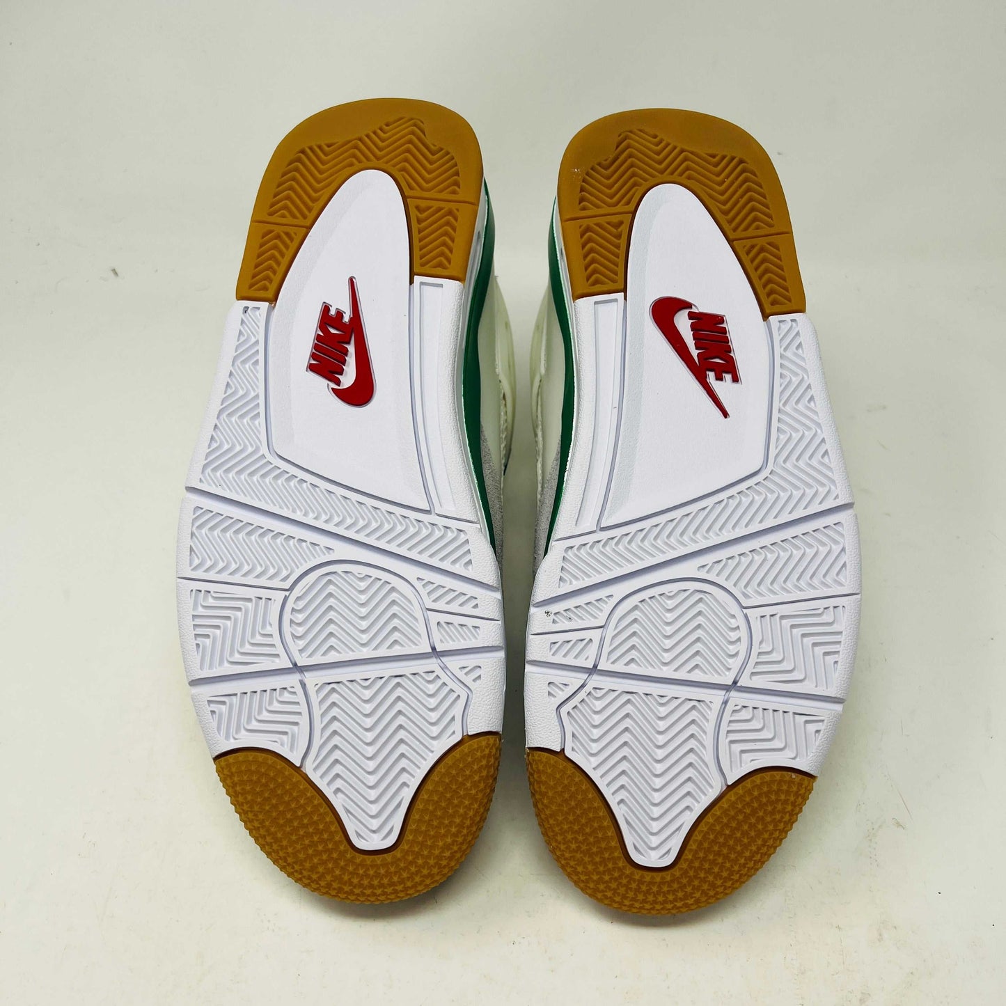 The image displays the soles of the Jordan 4 SB Pine Green shoes by Jordan. They have a predominantly white color with a patterned grip, featuring brown accents at the toe and heel. Each sole showcases a prominent red Nike logo.