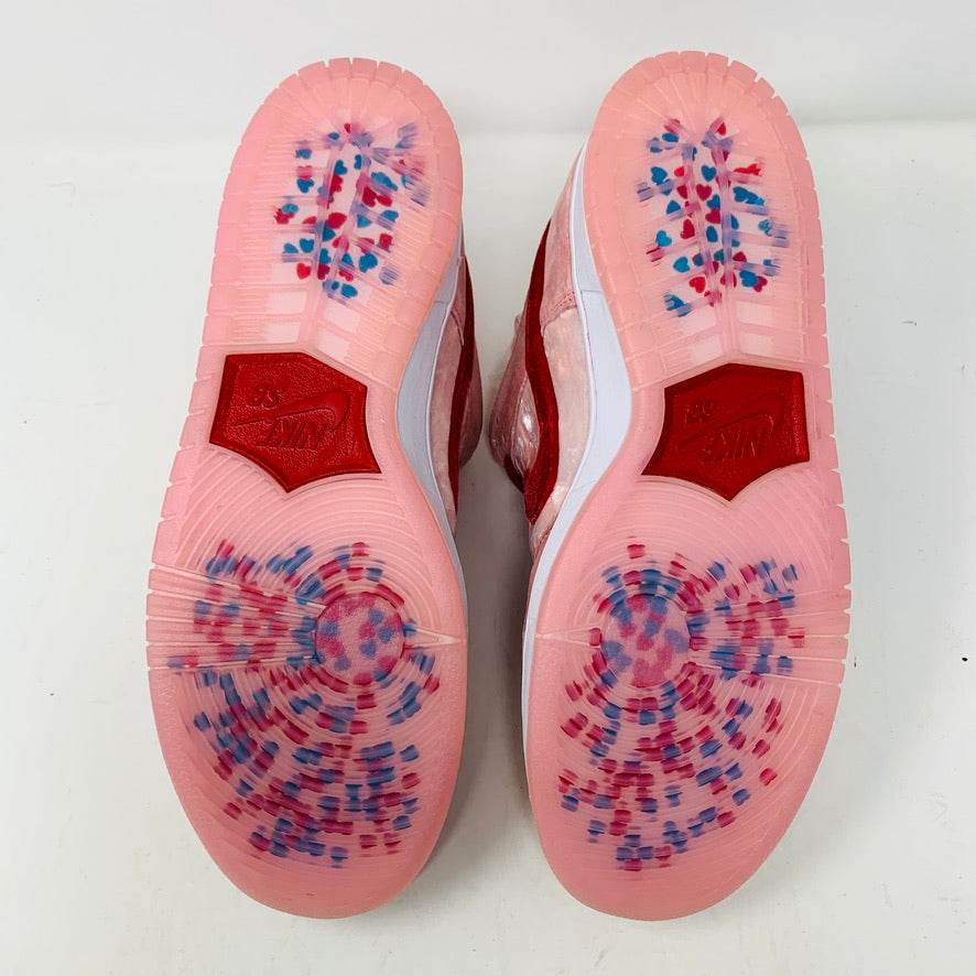 The Nike SB Dunk Low StrangeLove Skateboards, with vibrant pink and red, features a bold red swoosh, matching laces, and cartoon heart graphics on the heel. Displayed on a teal box with extra laces included, their design stands out against a plain white backdrop.