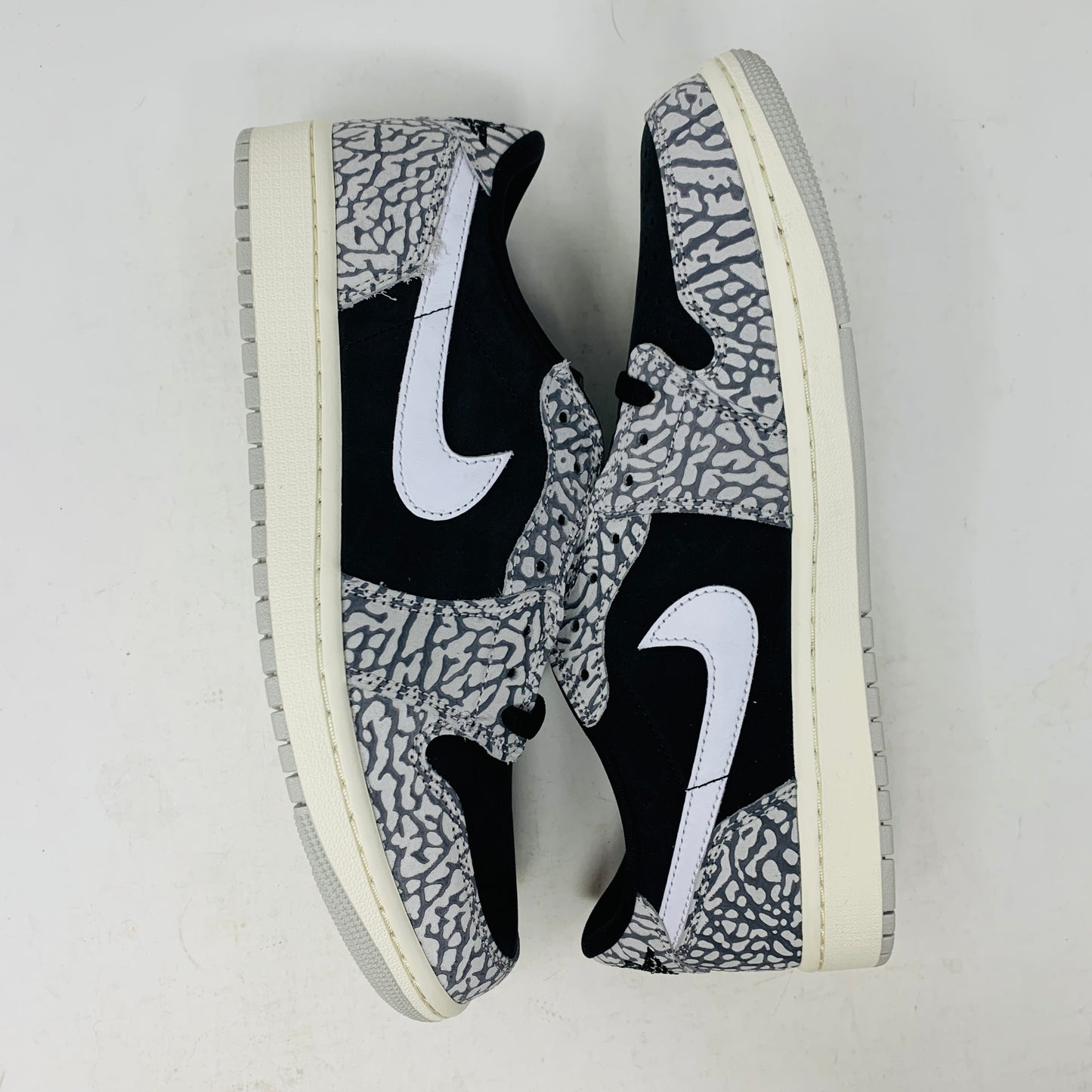 The Jordan 1 Retro Low OG Black Cement sneaker, with black and gray elephant print and a white swoosh, is showcased on a matching shoe box. A small brown box with extra laces is beside it, all set against a plain background.