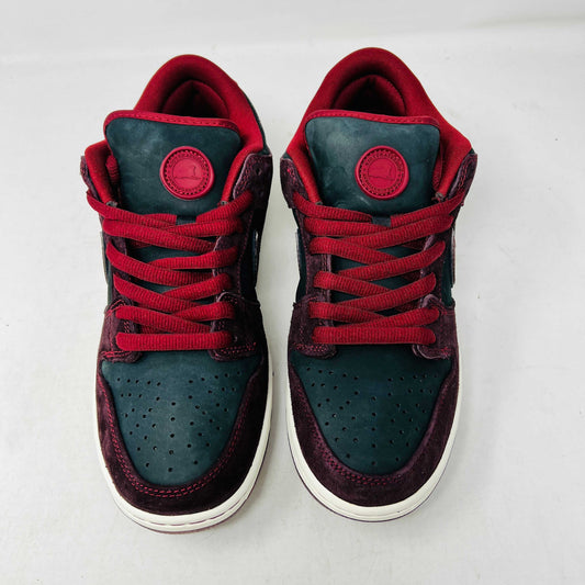 A BRAND NEW Nike SB Dunk Low Riot Skateshop sneaker featuring a black design with maroon accents, including a swoosh, laces, and a black sole with a white midsole, is displayed on top of its original white Nike SB shoebox.