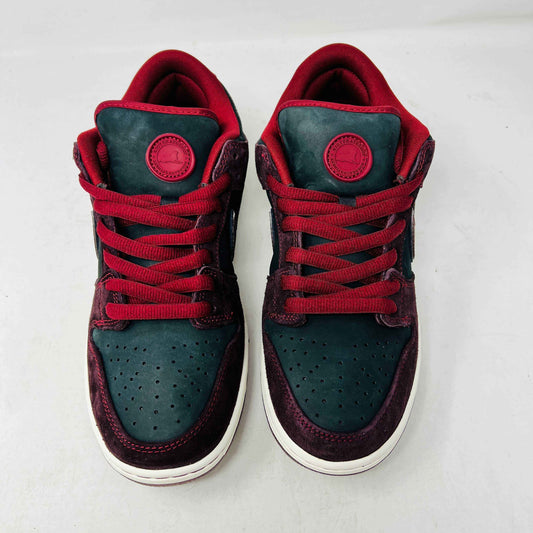 A BRAND NEW Nike SB Dunk Low Riot Skateshop sneaker featuring a black design with maroon accents, including a swoosh, laces, and a black sole with a white midsole, is displayed on top of its original white Nike SB shoebox.