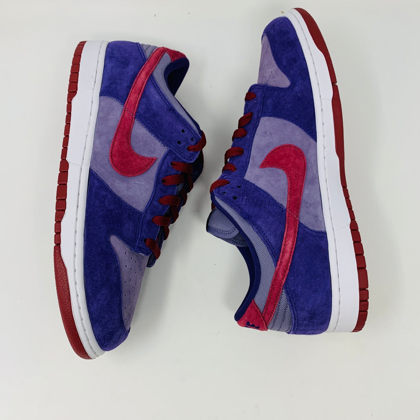 A pair of Nike Dunk Low Plum sneakers, in purple and maroon, rests on a red shoe box featuring a white Nike logo on a plain background. The shoes showcase a red swoosh and white midsoles, confirming their authenticity from Nike.