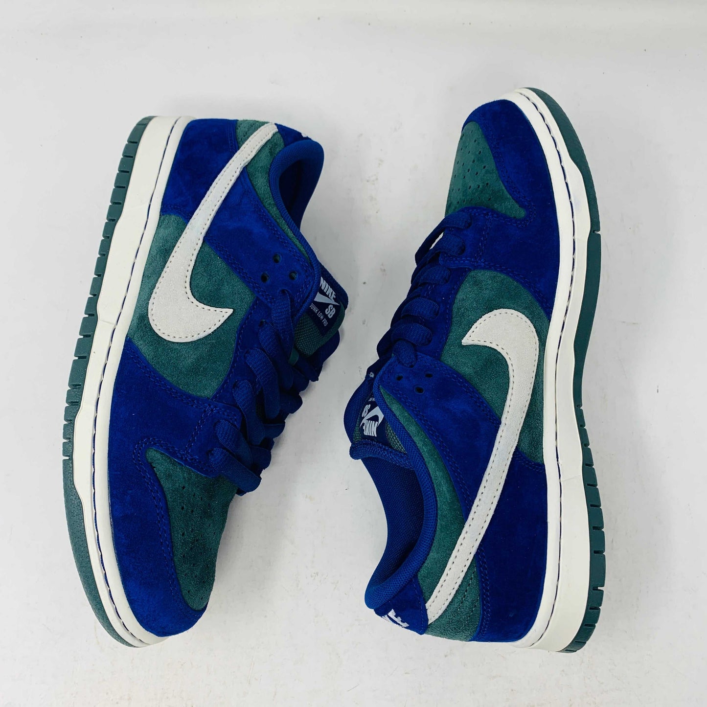 The Nike SB Dunk Low Deep Royal Blue sneaker, featuring a sleek blue and green design with a white swoosh, sits atop a purple Nike box. A bonus set of green laces accompanies the shoe, perfect for updating your style in 2024.