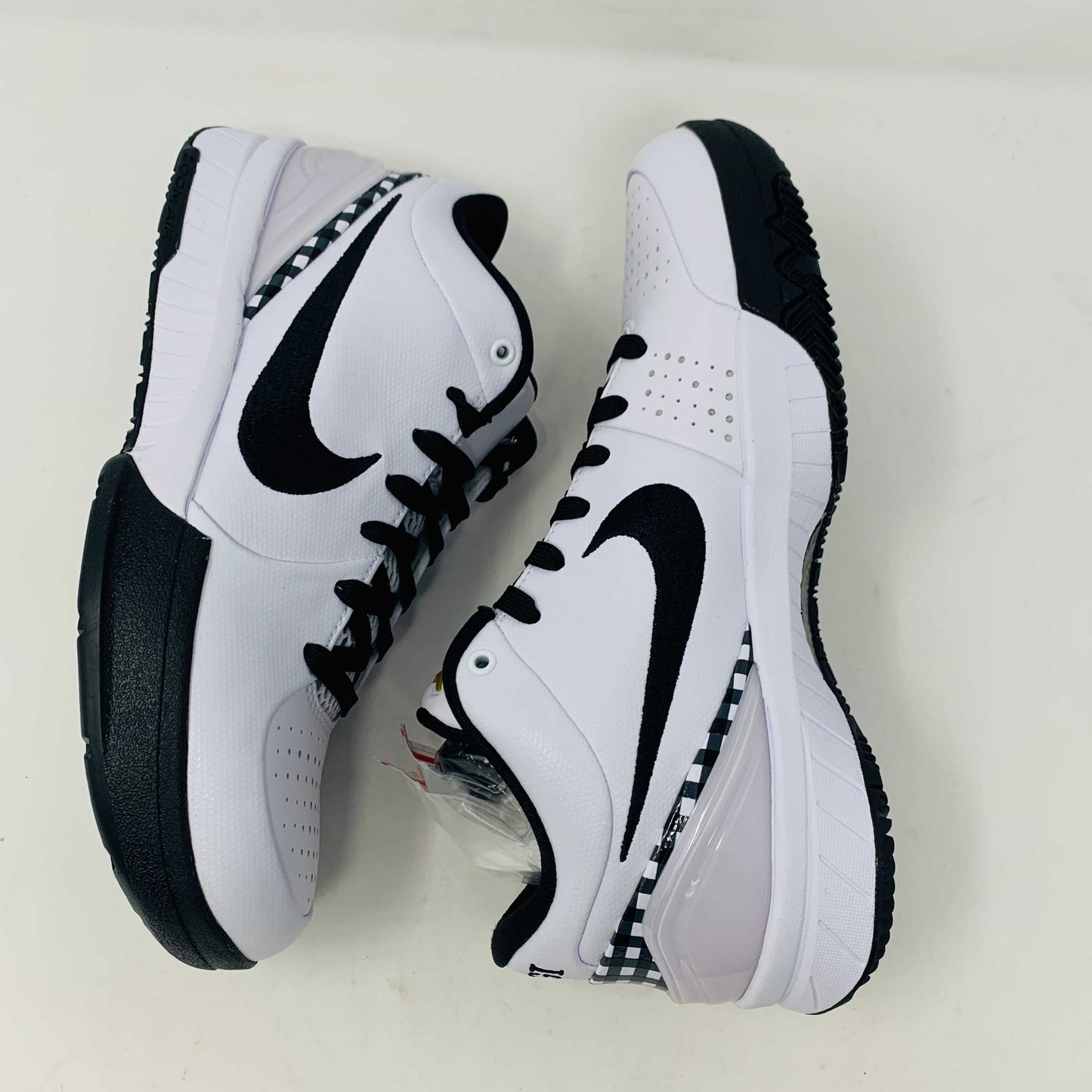 The Nike Kobe 4 Protro Mambacita Gigi shoe, featuring a black swoosh, black accents, and a checkered heel pattern, is displayed on a white box with a gold Nike logo. The box also includes extra laces.