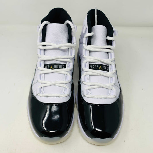 The Jordan 11 Retro DMP Gratitude (2023) features high-top sneakers in a white-black colorway with shiny accents. They have white laces, a crisp white midsole, and a translucent outsole, adorned with a small golden ankle logo and come in a sleek black box.