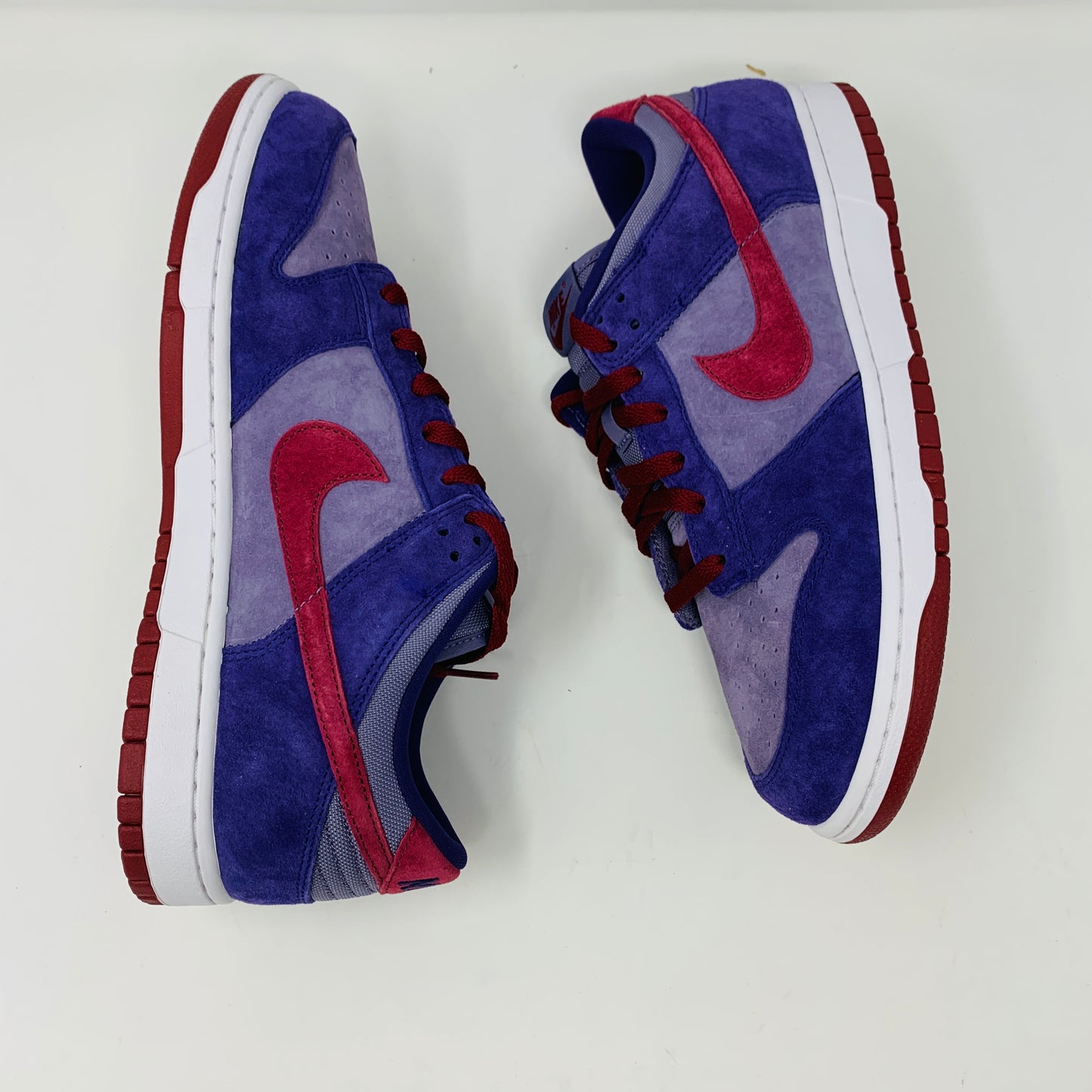 A pair of Nike Dunk Low Plum sneakers, in purple and maroon, rests on a red shoe box featuring a white Nike logo on a plain background. The shoes showcase a red swoosh and white midsoles, confirming their authenticity from Nike.