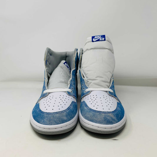 The Jordan 1 Hyper Royal, a high-top sneaker in white and light blue with a gray Nike swoosh and iconic Air Jordan logo, is showcased on a black and red Nike shoebox. It includes extra laces and features a crisp white midsole.