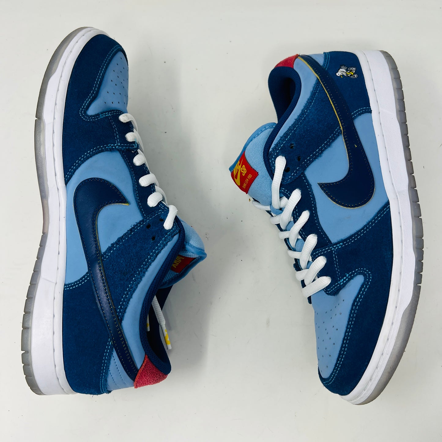 A Nike SB Dunk Low Pro Why So Sad? shoe, with blue uppers and white sole, rests on a purple box. It features an embroidered graphic on the heel, a dark blue swoosh, and extra laces in front. Price tags from 2022 are visible beside it.