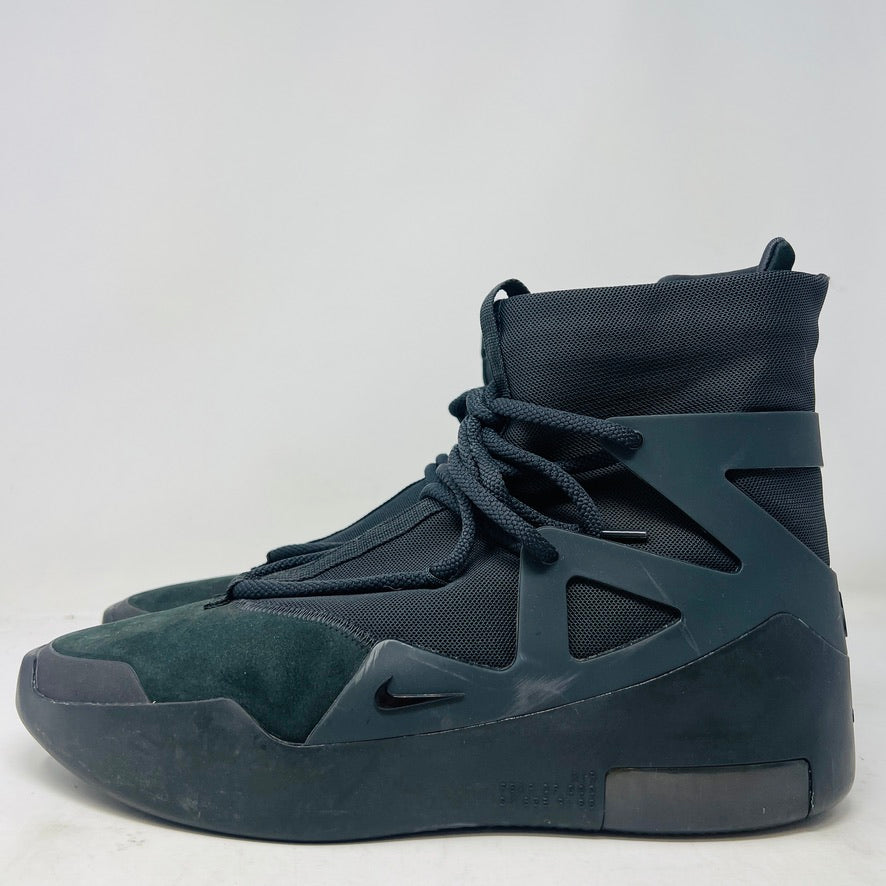 The Nike Air Fear of God 1 Triple Black is a high-top, dark, minimalist sneaker with angular overlays and a textured sole. It features a high collar, matte finish, dark laces, and has a small logo near the midsole. The background is light gray with general wear signs.
