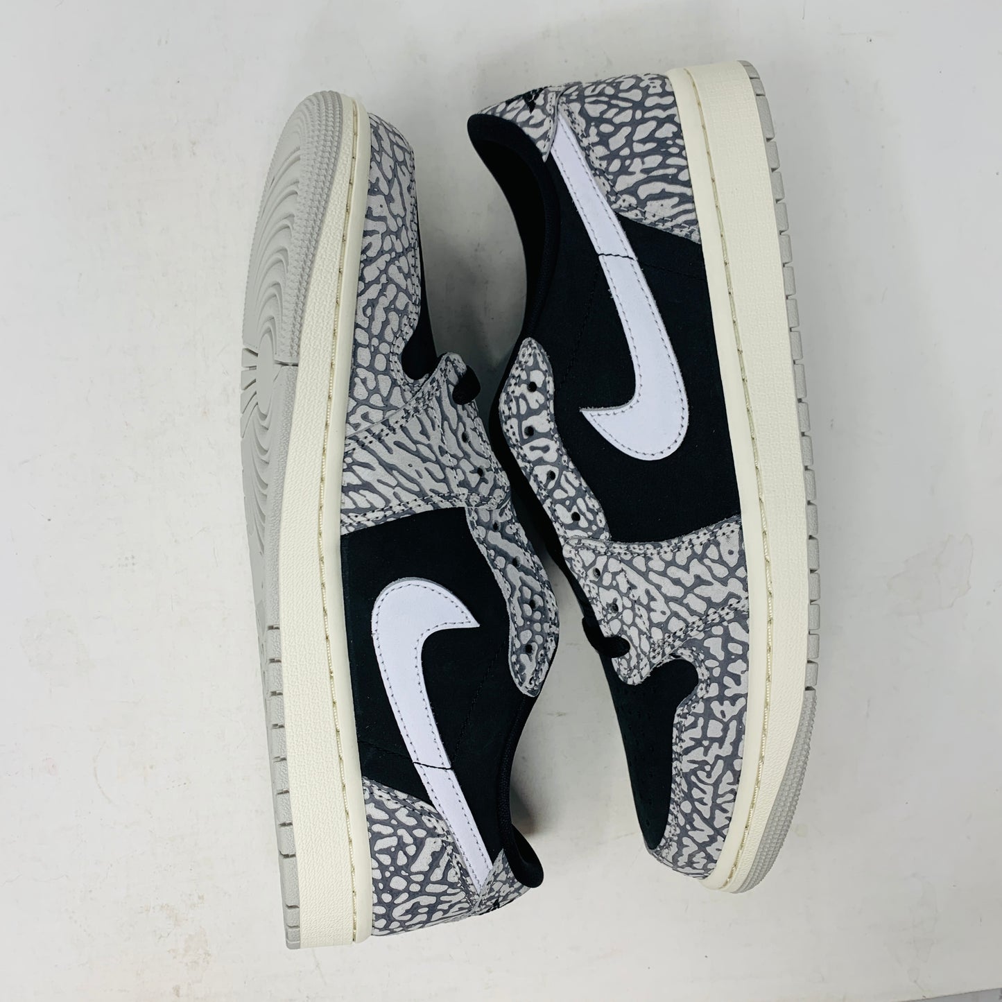 The Jordan 1 Retro Low OG Black Cement sneaker, with black and gray elephant print and a white swoosh, is showcased on a matching shoe box. A small brown box with extra laces is beside it, all set against a plain background.