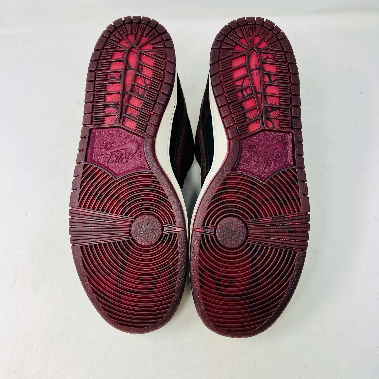 A BRAND NEW Nike SB Dunk Low Riot Skateshop sneaker featuring a black design with maroon accents, including a swoosh, laces, and a black sole with a white midsole, is displayed on top of its original white Nike SB shoebox.