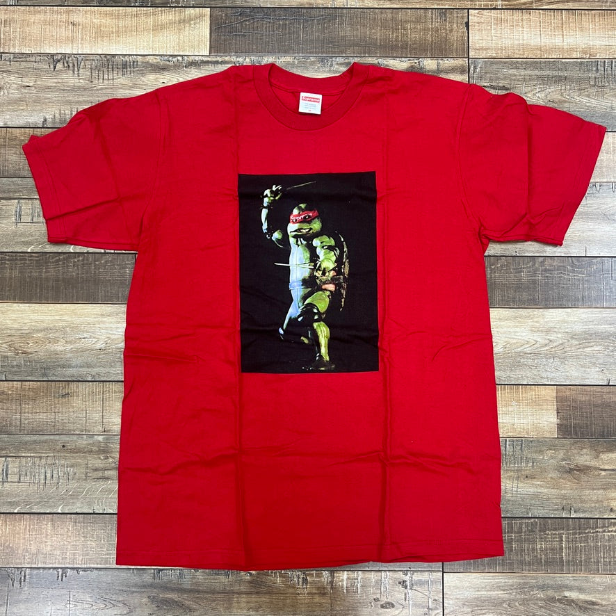 The Supreme Raphael Tee Red by Supreme, a 100% authentic red t-shirt, showcases an anthropomorphic turtle in red with sais on a black backdrop. It makes a bold statement while laid flat on a wooden floor.