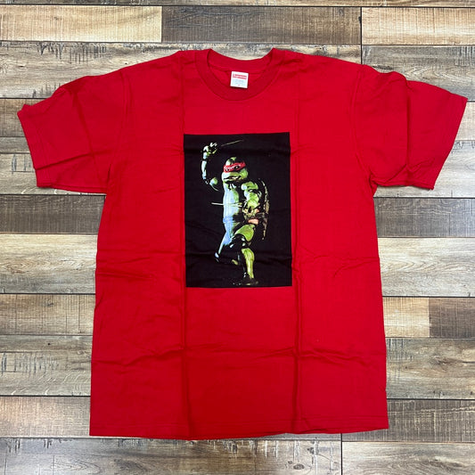 The Supreme Raphael Tee Red by Supreme, a 100% authentic red t-shirt, showcases an anthropomorphic turtle in red with sais on a black backdrop. It makes a bold statement while laid flat on a wooden floor.